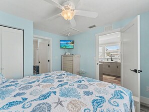 Ein Schlafzimmer mit einem Bett, einem Deckenventilator, einem Fernseher, einer Kommode und einem durch eine offene Tür einsehbarer Badezimmer.