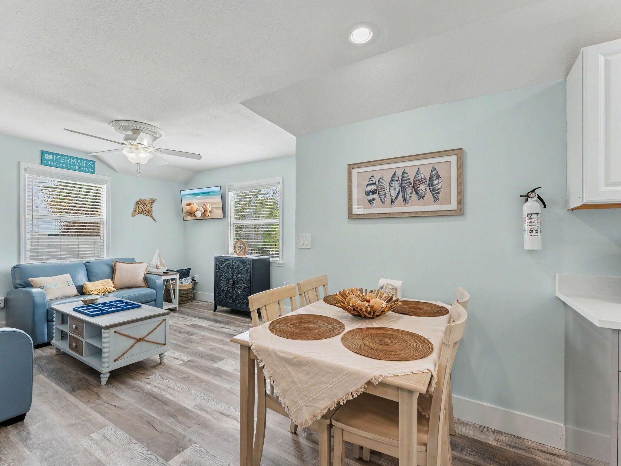 A living room with a blue sofa, a wooden dining table with woven placemats, a ceiling fan, and a framed seashell artwork on the wall.