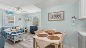 A living room with a blue sofa, a wooden dining table with woven placemats, a ceiling fan, and a framed seashell artwork on the wall. - Sunset Paradise (Madeira Beach)