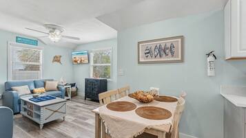 A living room with a blue sofa, a wooden dining table with woven placemats, a ceiling fan, and a framed seashell artwork on the wall.