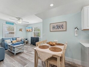 Ein Wohnzimmer mit einem blauen Sofa, einem Holzesstisch mit gewebten Tischsets, einem Deckenventilator und einer gerahmten Wandkunst mit Meeresschnecken.
