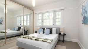 Quarto com um espelho grande, uma cama com manta xadrez, duas mesas de cabeceira com abajures e uma janela com vitrais.