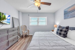 Une chambre à coucher comprenant un lit, une commode, une chaise, un ventilateur de plafond, une fenêtre avec des stores et un tableau accroché au mur.