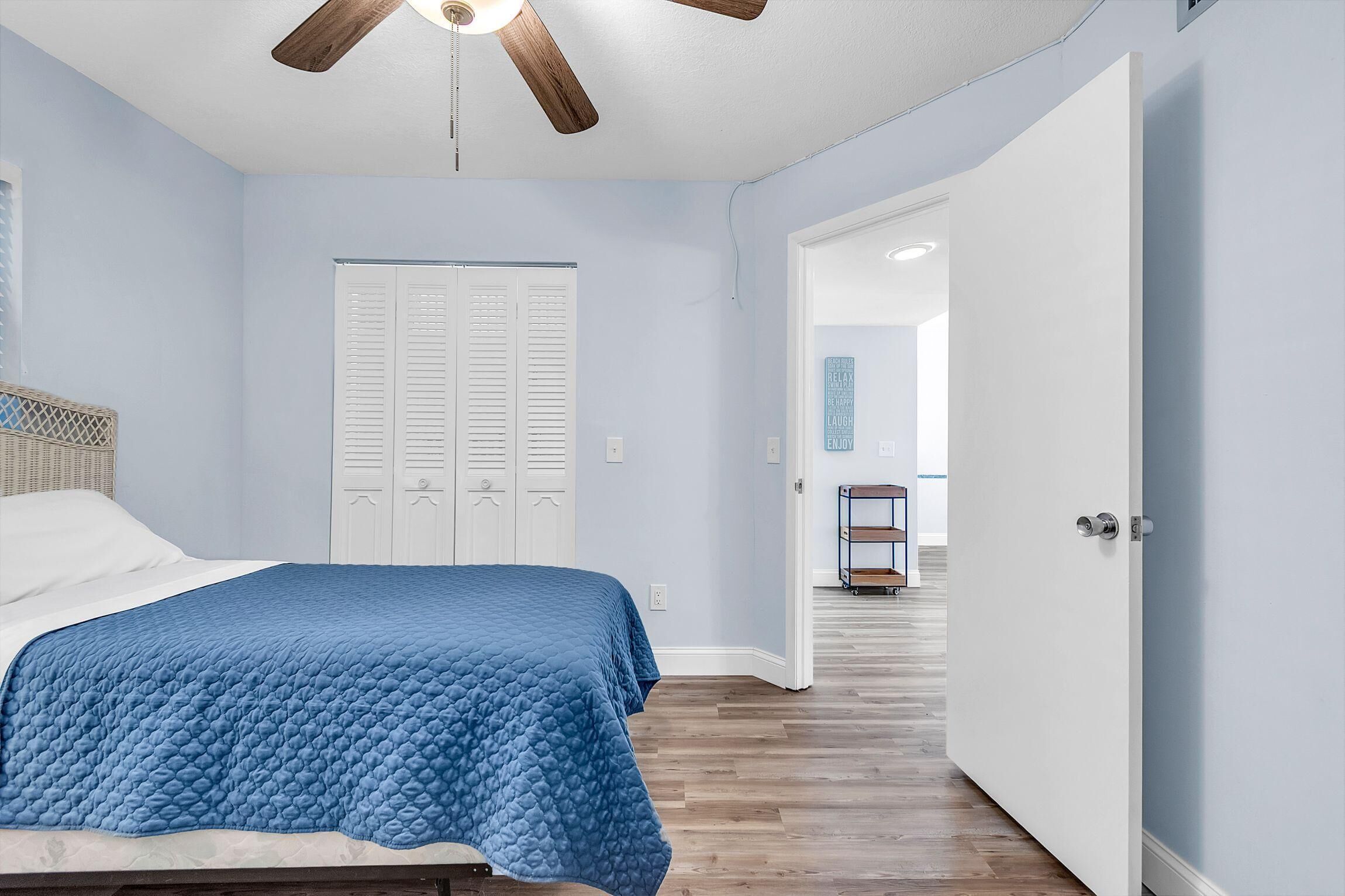 A bedroom with a bed, a ceiling fan, a white door, and a hallway leading to another room.