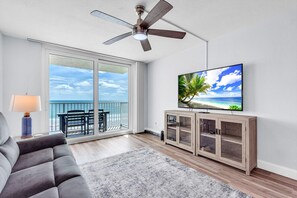 A living room with a ceiling fan, a TV on a wooden cabinet, a sofa, a lamp, and a view of the ocean through large sliding doors. - Gulf Shores #402 (Indian Shores)