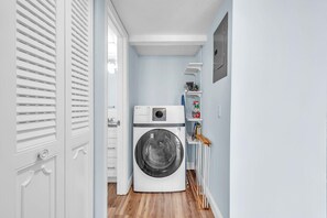 Une buanderie équipée d’un lave-linge à chargement frontal, d’une étagère contenant des produits de nettoyage et d’une porte à persiennes.