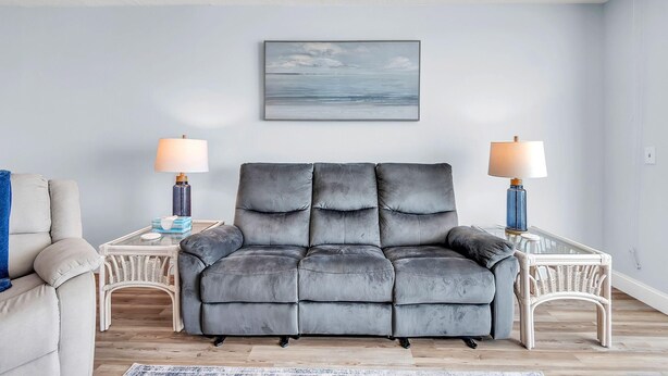 Ein graues Sofa mit passendem Hocker, zwei Beistelltische mit Lampen, ein Korbstuhl und ein gerahmtes Kunstwerk an der Wand.