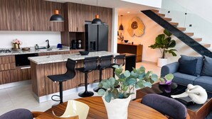 Et moderne køkken med træskabe, en central ø med barstole, et spiseområde med en sofa og en unik skulptur, samt en trappe med gelænder af glas.