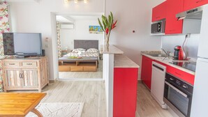 Cocina pequeña con armarios rojos, lavavajillas y microondas. Se ve una cama con colcha estampada al fondo.