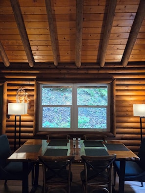 Interior de uma cabana de madeira com mesa de jantar, cadeiras, janela com vista para a vegetação e um abajur.
