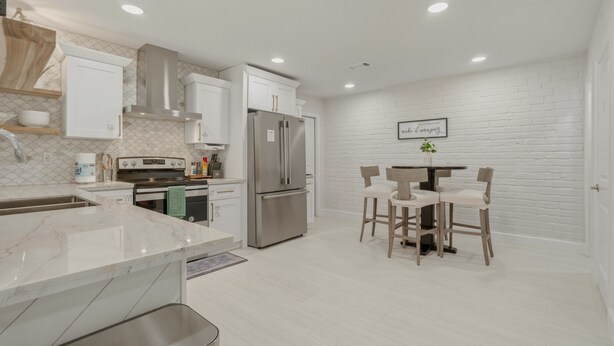 A modern kitchen with white cabinetry, a stainless steel refrigerator, and a dining area with wooden chairs. - Bean House Near NASA • Kemah Boardwalk • Galveston (Webster)