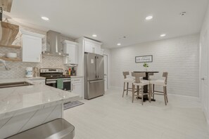 A modern kitchen with white cabinetry, a stainless steel refrigerator, and a dining area with wooden chairs. - Bean House Near NASA • Kemah Boardwalk • Galveston (Webster)