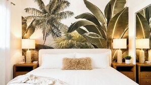Una cama bien hecha con un almohadón esponjoso, una lámpara de noche y una mesita de noche con una planta pequeña.