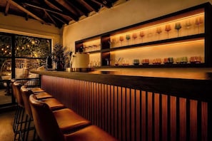 Un espace bar avec un comptoir et des tabourets en bois, des étagères remplies de verres et une vue sur la verdure à l’extérieur.