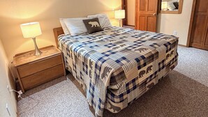 A bedroom with a bed, two bedside lamps, a wooden nightstand, a carpeted floor, and a wooden door.