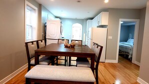 Un comedor con una mesa y bancos de madera, una cocina con electrodomésticos blancos, y un dormitorio visible a través de una puerta abierta.