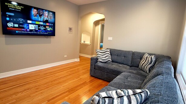 Una sala de estar moderna con un sofá modular, piso de madera, un televisor montado que muestra un servicio de streaming y un ventilador de techo.