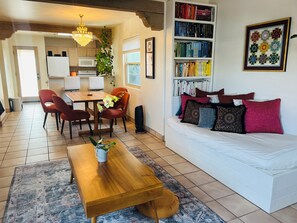 Ein Wohnzimmer mit Couchtisch aus Holz, einem weißen Sofa mit gemusterten Kissen, einem Bücherregal und einem Essbereich mit Stühlen und Tisch.