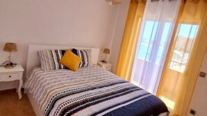 Un dormitorio con una cama, dos mesitas de noche, una lámpara y un ventanal con cortinas.