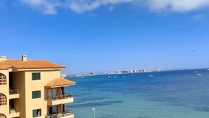 Un edificio de varios pisos con balcones que dan a un mar azul y despejado, un muelle a lo lejos y un cielo claro.