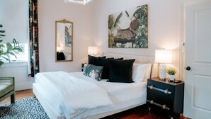 Un dormitorio con cama, mesitas de noche, un espejo, un cuadro y una puerta.