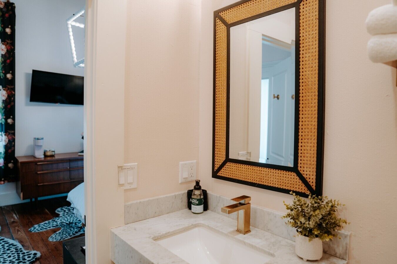 A bathroom with a large mirror, a sink with a gold faucet, a bottle of hand soap, and a potted plant.