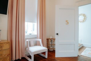 Una habitación con una puerta blanca marcada con el número 5, una silla blanca, un aparador de madera y una ventana con cortinas.
