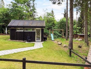 A small wooden cabin with a metal roof, a patio with a chair, and a swing set in a forested area. - 5 Star Holiday Home in Ans By (Ans)