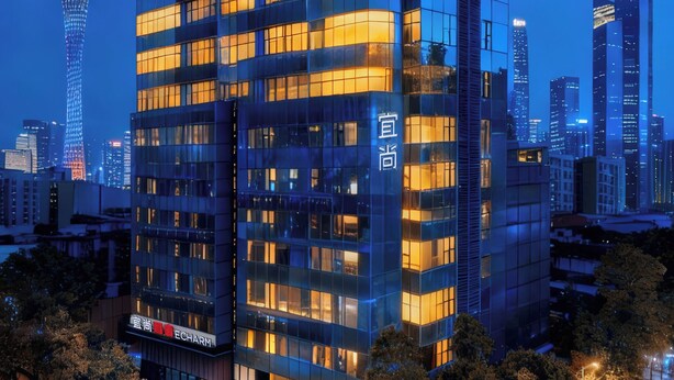 Un moderno edificio de cristal con ventanas iluminadas, rodeado de árboles y el horizonte de una ciudad de noche.