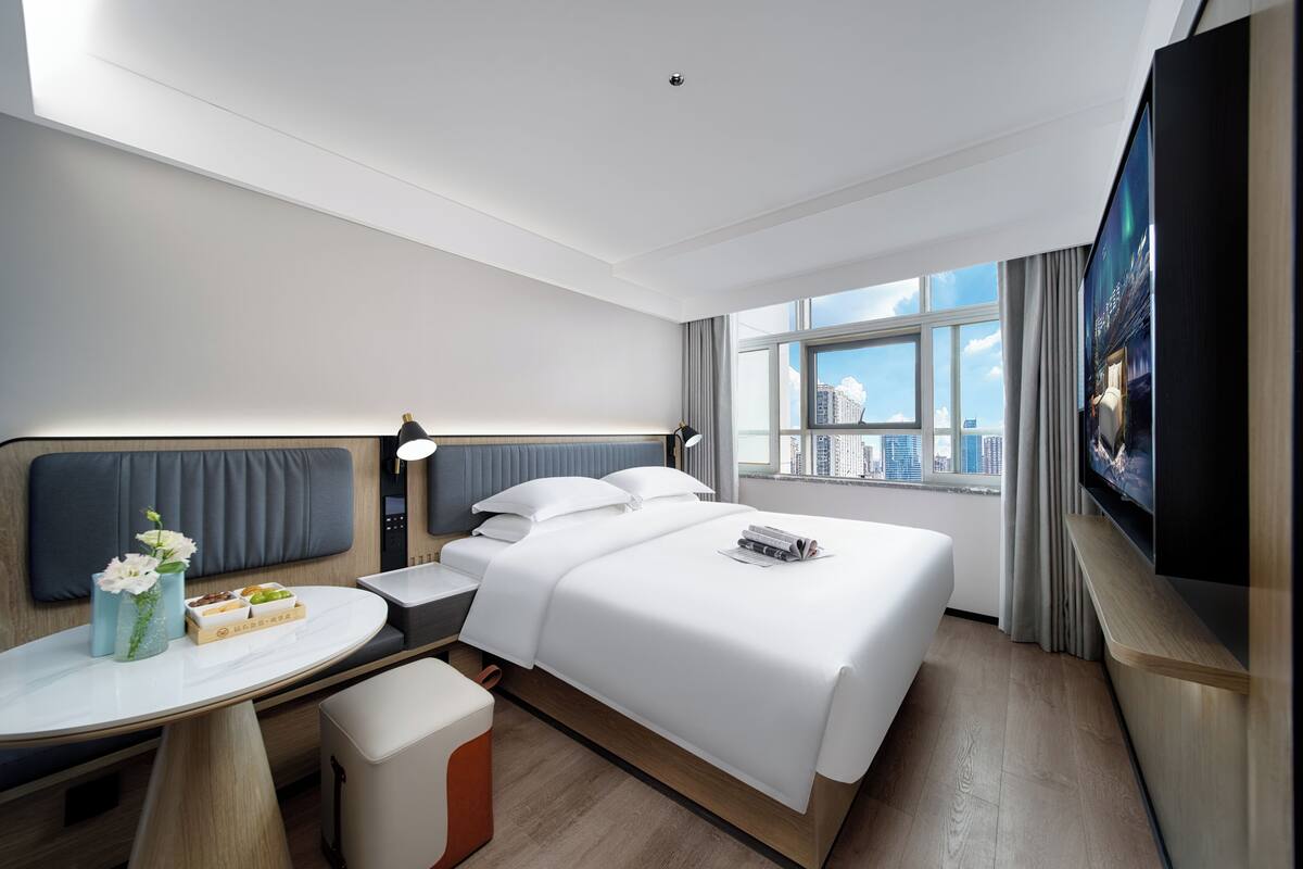 A hotel room with a bed, bedside table, TV, and a window with a city view.