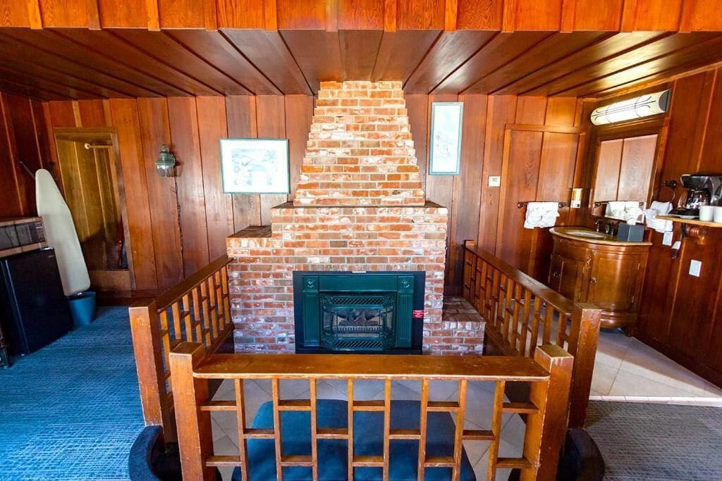 A wooden interior with a brick fireplace, wooden benches, and a kitchen area.
