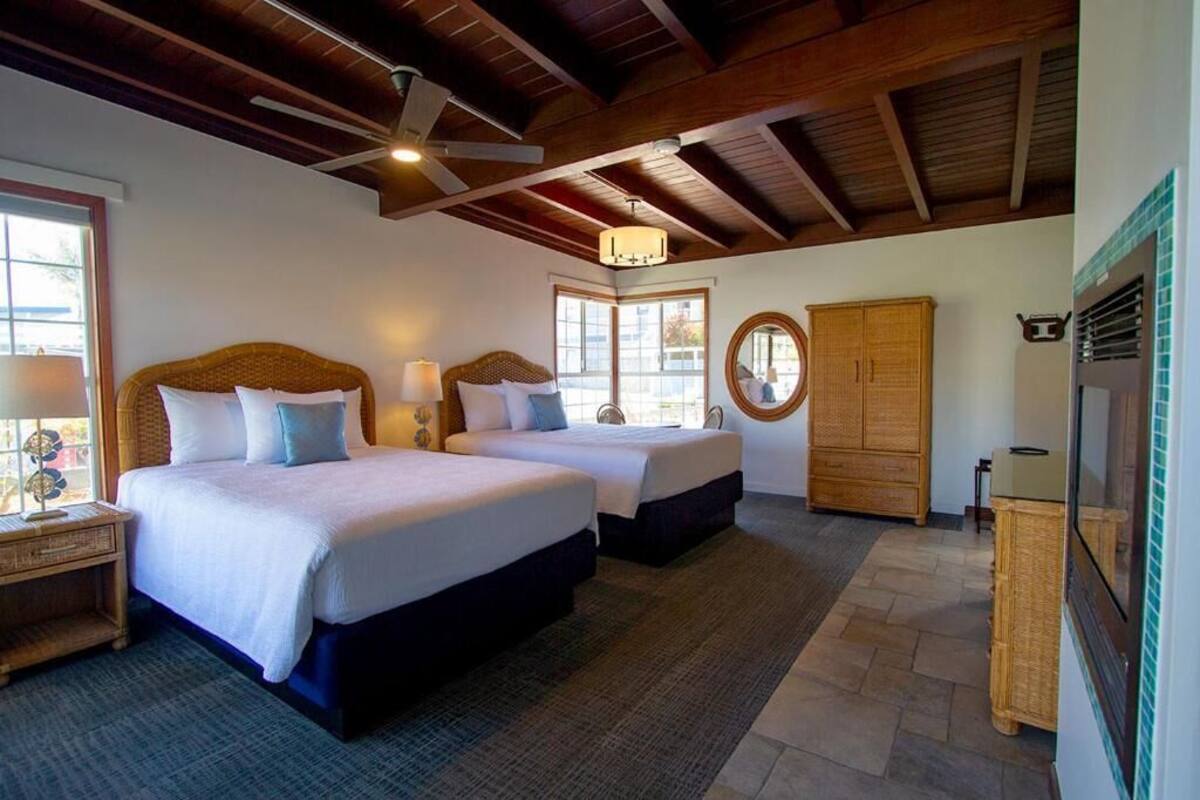 A hotel room with two beds, a ceiling fan, a large window, and a wooden wardrobe.