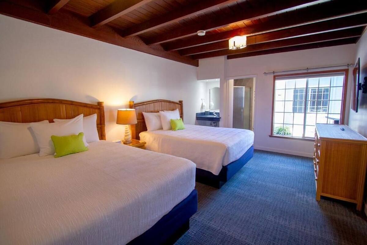 A hotel room with two beds, a wooden dresser, a bathroom with a mirror, and a large window.