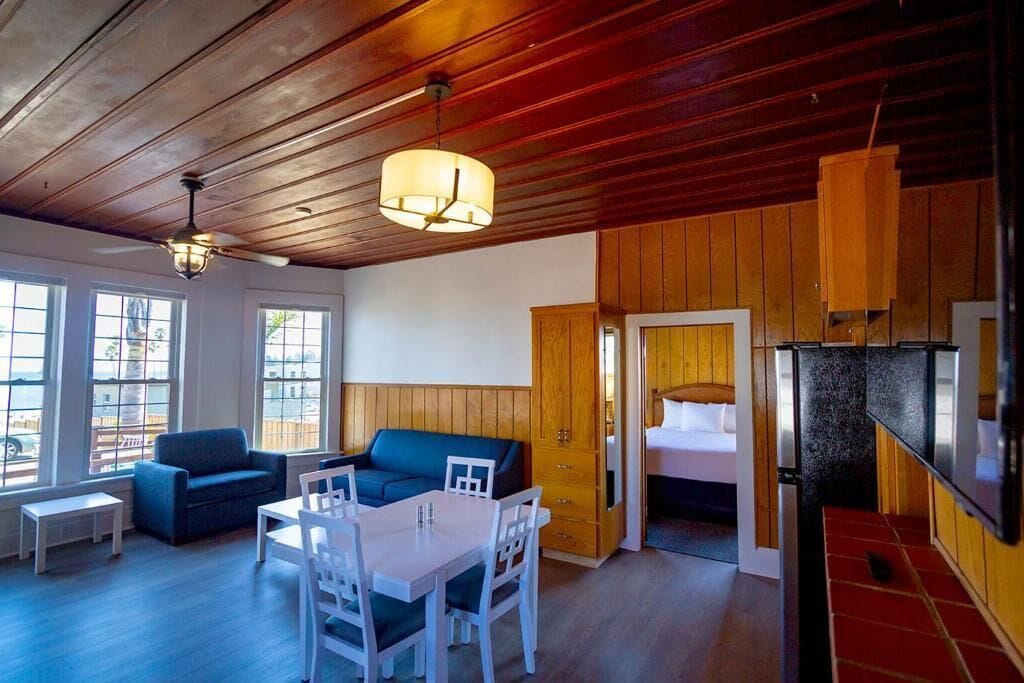 A room with a wooden ceiling, a dining table with chairs, a sofa, and a bed with a wooden door.