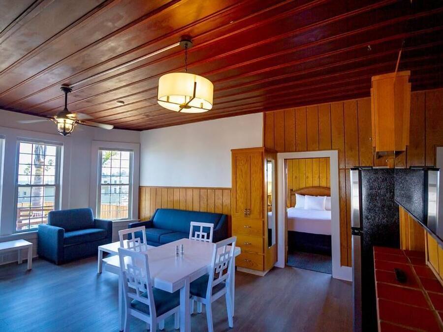 A room with a wooden ceiling, a dining table with chairs, a sofa, and a bed with a wooden door.