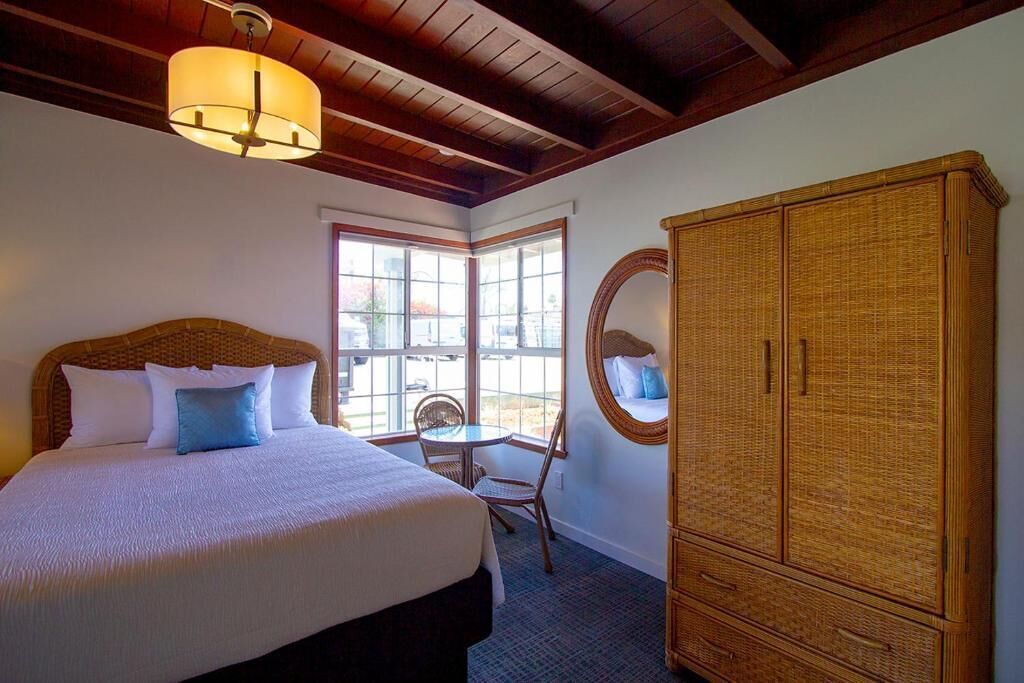 Una habitación de hotel con una cama grande, un espejo redondo, un armario de mimbre y una pequeña mesa con sillas.