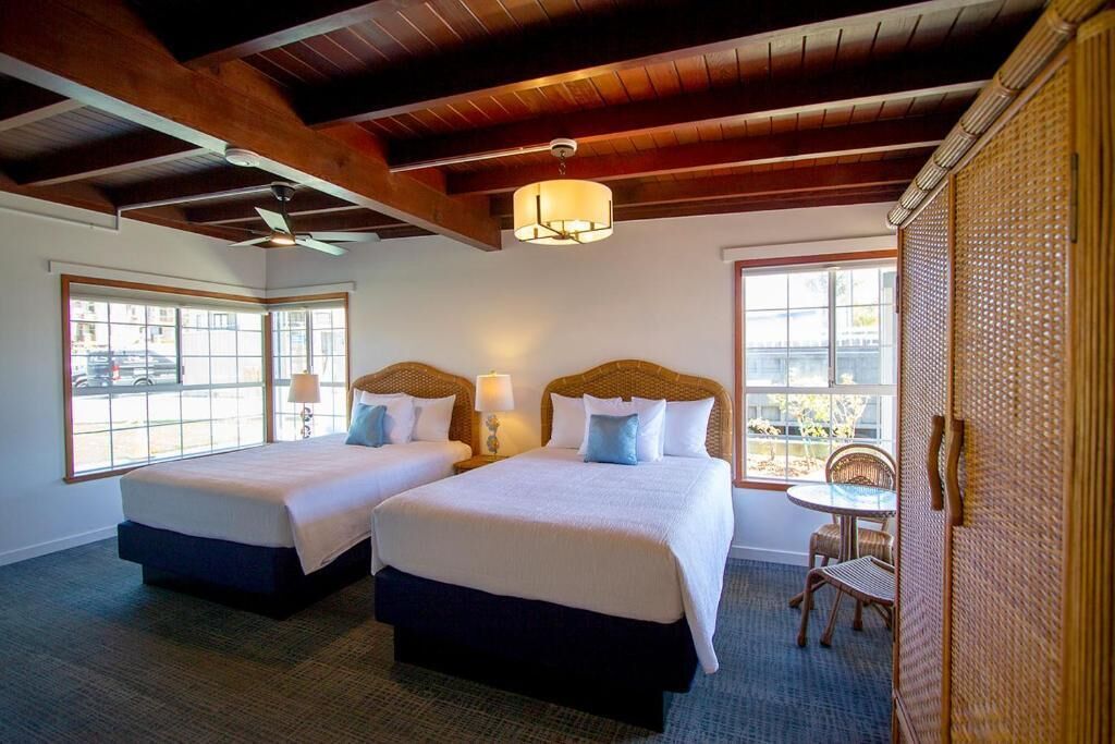 Habitación de hotel con dos camas, ventilador de techo, una mesa con sillas y ventanas amplias.