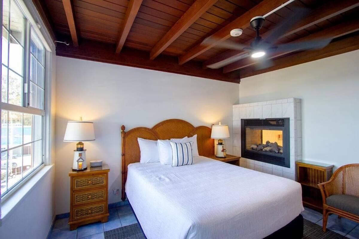 A bedroom with a wooden headboard, two bedside lamps, a fireplace, a window, and a chair.