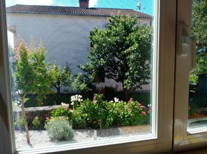 A window view of a building with a tiled roof, a garden with various plants and flowers, and a clear sky. - One Bedroom Apartment with Terrace (Rovinj)