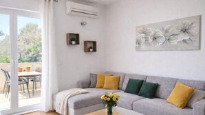 Uma sala de estar com um sofá cinza, almofadas amarelas e verdes, uma mesa de centro de madeira e um vaso de flores. Há uma porta de vidro deslizante que dá para uma área externa com uma mesa e cadeiras.