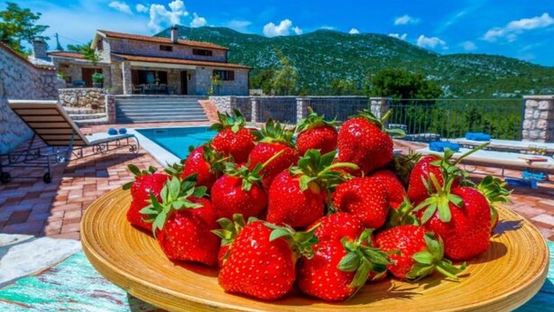 A poolside area with a plate of strawberries, a stone house, and a mountain in the background. - Villa with swimming pool (Vrgorac)