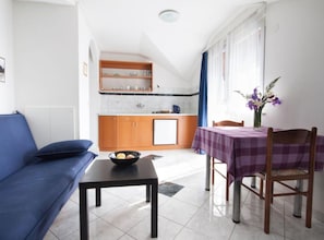 A living room with a sofa, a dining table with chairs, a kitchen area, and a window with curtains. - One Bedroom Apartment with small Balcony (Umag)