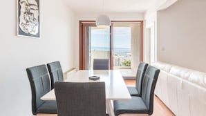 Un comedor moderno con una mesa blanca y cuatro sillas grises, una lámpara colgante y vistas al mar a través de un ventanal grande.