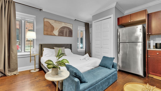 Ein modernes Hotelzimmer mit Bett, blauem Sofa, Küchenzeile inklusive Kühlschrank und Fenster mit Vorhängen.
