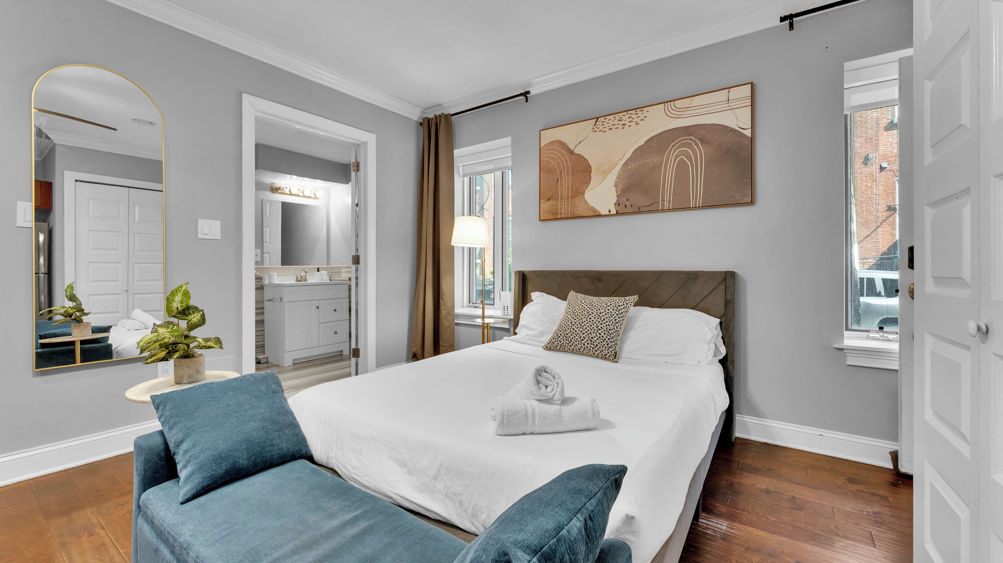 Habitación de hotel con cama, mesita de noche con lámpara, un espejo grande, baño visible a través de una puerta abierta y un cuadro en la pared.