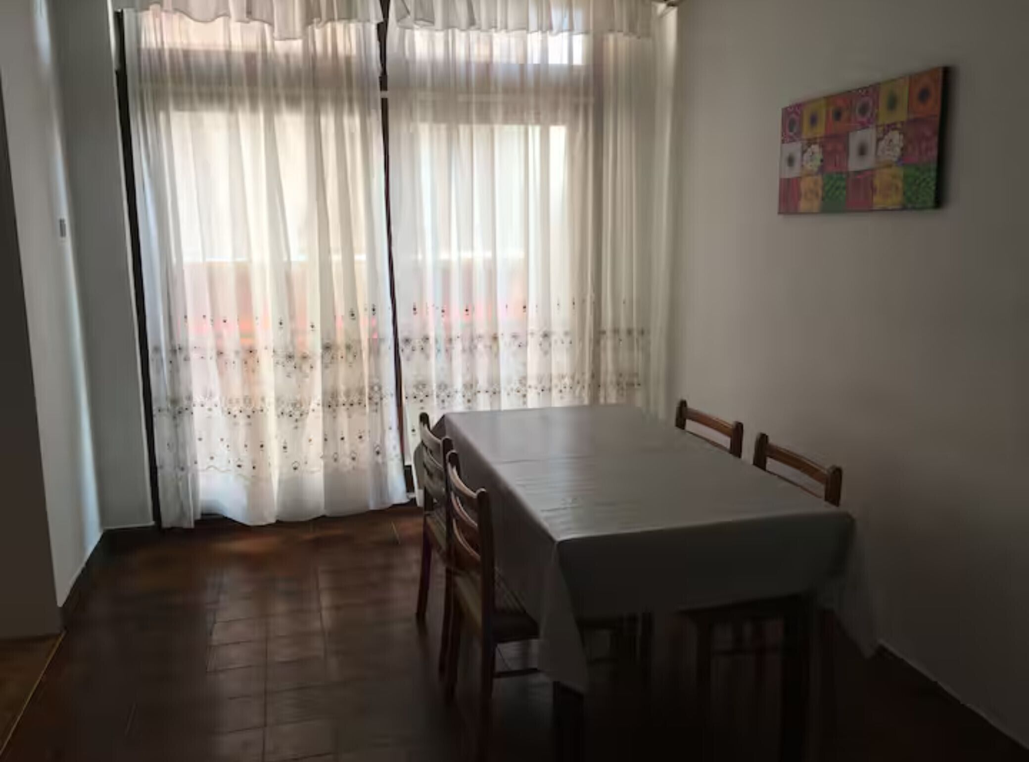 A dining area with a table and chairs, a window with sheer curtains, and a colorful artwork on the wall.