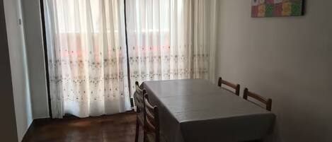 A dining area with a table and chairs, a window with sheer curtains, and a colorful artwork on the wall.