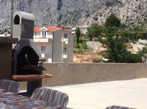 A patio with a grill, a table with a colorful tablecloth, and a view of a mountain and buildings. - Three Bedroom Apartment on Ground floor (Drasnice)