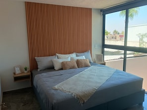 Ein modernes Schlafzimmer mit einem großen Bett, einem Holzschrank als Kopfteil, einem Nachttisch mit Pflanze und Blick ins Freie.