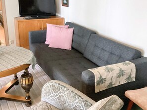 A living room with a grey sofa, a wooden cabinet, a television, and a patterned throw blanket. - 5 Person Holiday Home in Bastad (Bastad)